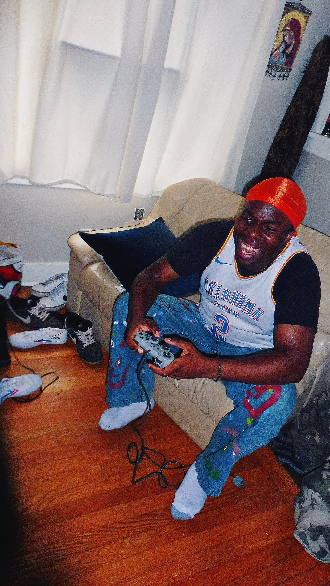 King E.D.O seated on a couch holding a game controller, wearing an orange durag and an Oklahoma City jersey.