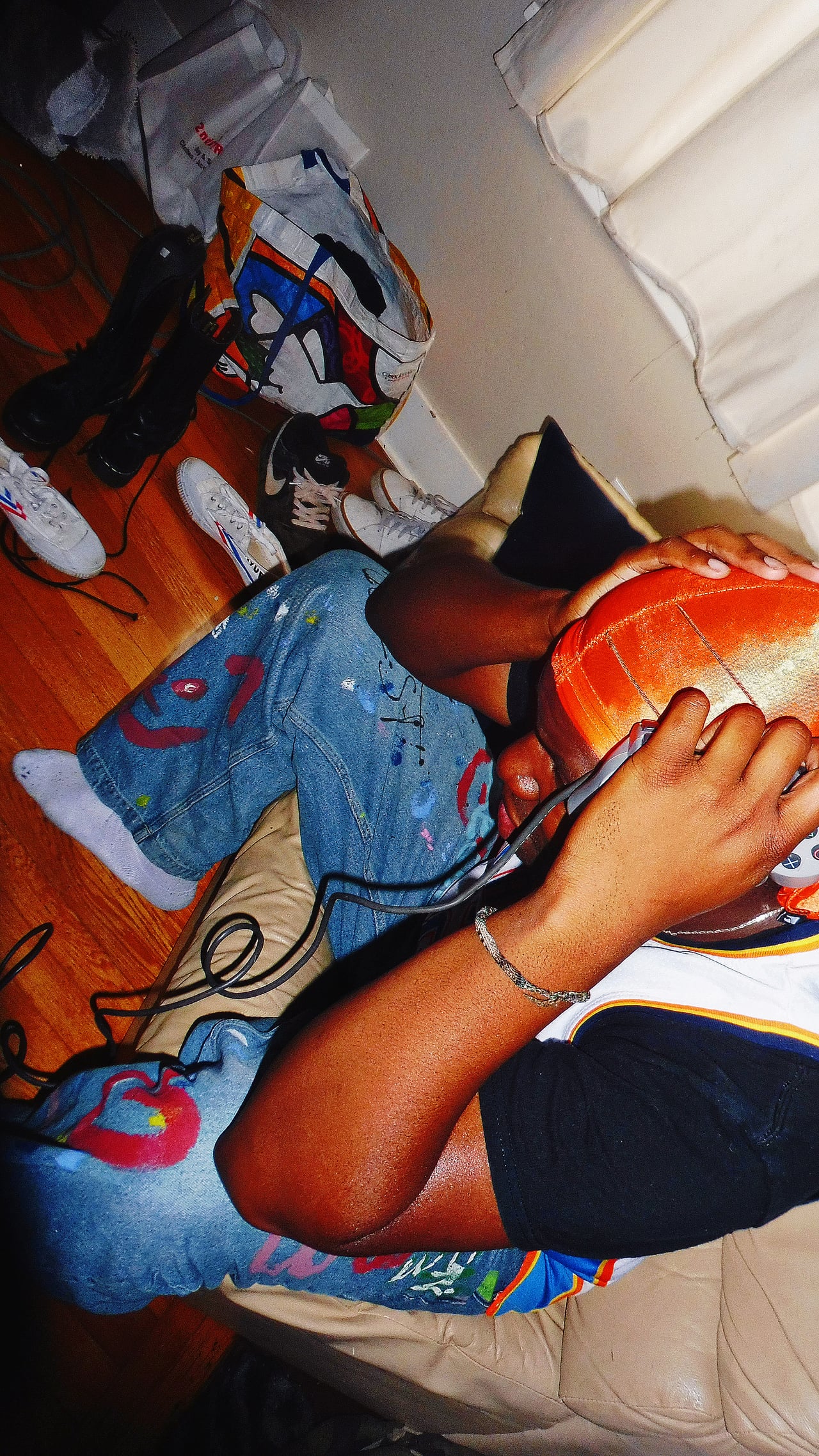 King E.D.O wearing an orange durag and holding headphones while seated on a couch, with a game controller beside him.