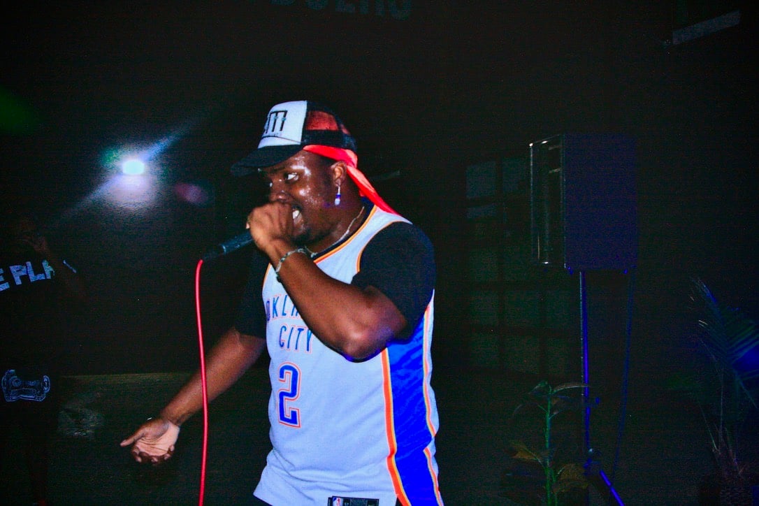 King E.D.O performing with a microphone, wearing an Oklahoma City jersey and a Burn Money cap.
