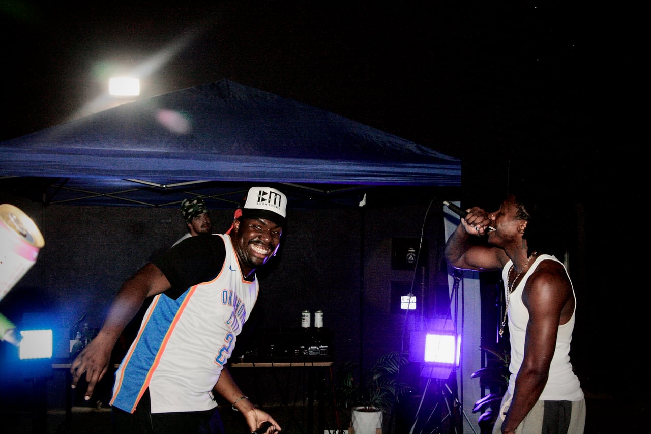 King E.D.O smiling during an outdoor live performance beneath a blue canopy, with another performer holding a microphone.